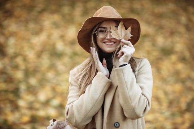 Immagine di una giovane donna attraente vestita con un cappotto e un cappello di feltro sullo sfondo delle foglie cadute nel parco in autunno