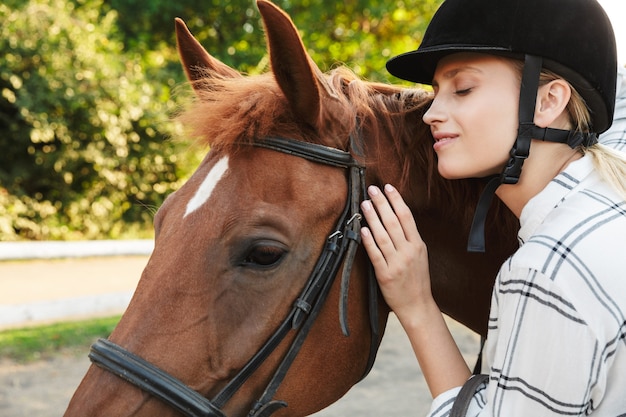 Immagine di una giovane donna bionda caucasica che indossa un cappello in piedi a cavallo in cantiere in campagna