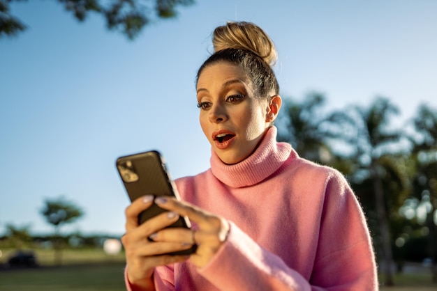 Immagine di una giovane donna bionda latina eccitata sorpresa utilizzando il cellulare sullo sfondo del parco