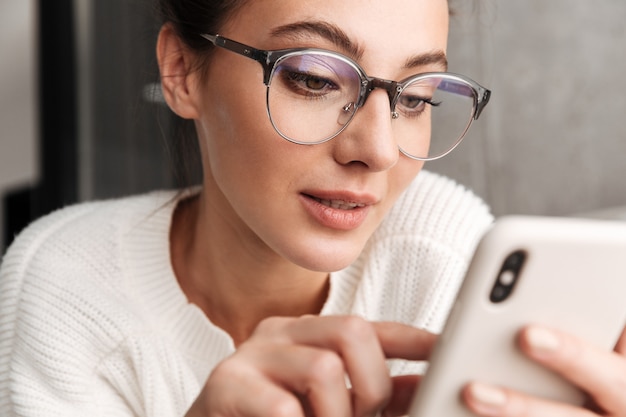 Immagine di una giovane donna bruna positiva con gli occhiali che sorride e tiene il cellulare a casa