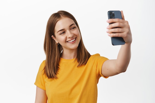 Immagine di una giovane donna che scatta foto sullo smartphone, posa per un selfie sul cellulare, sorride e sembra felice, parla su un'app per videochiamate, in piedi su sfondo bianco