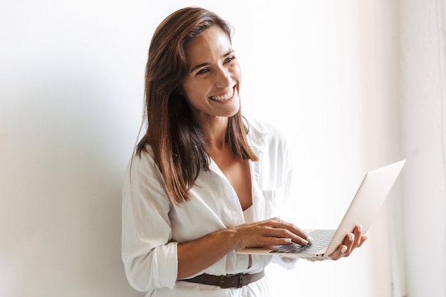 Immagine di una giovane donna d'affari positiva sul posto di lavoro al chiuso vicino alla finestra utilizzando il computer portatile.