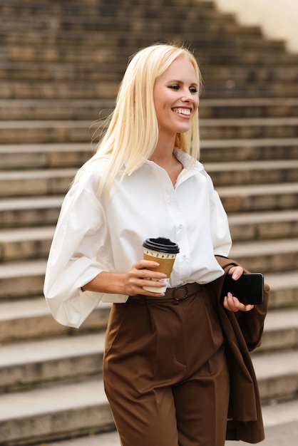 Immagine di una giovane donna d'affari sorprendente allegra felice che cammina per gradini all'aperto in abiti da cerimonia con caffè.