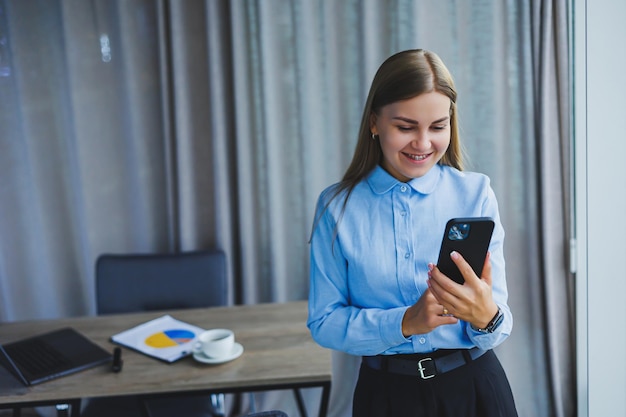 Immagine di una giovane donna felice con una giacca sorridente e al lavoro su un laptop che parla al telefono in un ufficio moderno con grandi finestre Lavoro a distanza