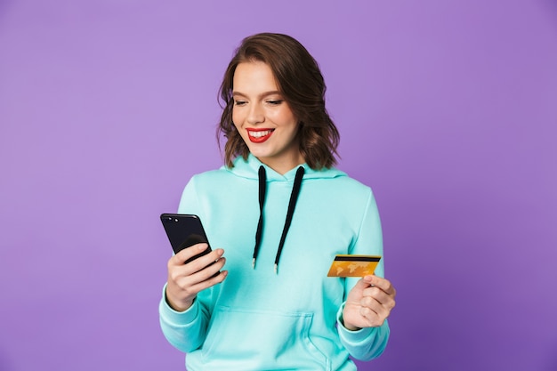 Immagine di una giovane donna felice in posa isolata sopra la parete viola muro utilizzando il telefono cellulare in possesso di carta di credito.