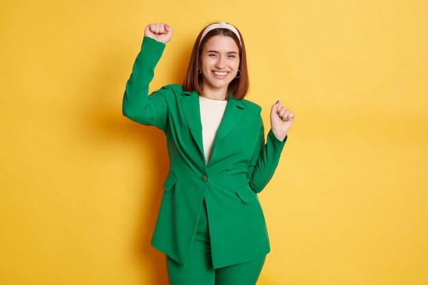 Immagine di una giovane donna sorridente felice che indossa un'elegante giacca verde in posa isolata su sfondo giallo in piedi con le braccia alzate che celebra il suo successo o la sua vittoria esprimendo felicità