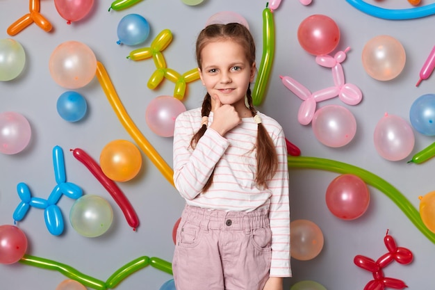 Immagine di una graziosa e affascinante bambina sorridente con le trecce che indossano abiti casual in posa isolate su sfondo grigio con palloncini che le tengono il mento mostrando il pollice in su