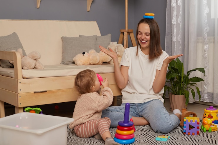 Foto immagine di una madre che ride positivamente indossando camicia bianca e jeans che gioca con la sua piccola figlia a casa mentre è seduta sul pavimento tenendo anelli di piramide sulla sua testa
