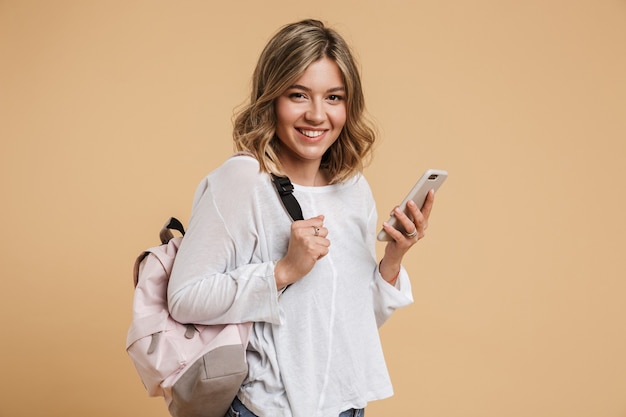 Immagine di una ragazza adolescente positiva che indossa uno zaino usando il cellulare e sorride isolata su un muro beige