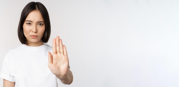 Immagine di una ragazza asiatica che mostra stop proibire smth estendere un braccio per mostrare il gesto proibito tabù in piedi in maglietta su sfondo bianco