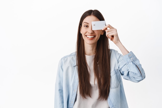 Immagine di una ragazza bruna sorridente, che mostra la carta di credito vicino al viso e sembra positiva, in piedi contro il muro bianco