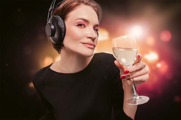 Immagine di una ragazza in abito nero con un bicchiere di vino in mano in una discoteca. Cuffie professionali. Concetto di festa. Tecnica mista
