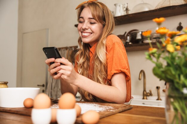 Immagine di uno chef di giovane donna bionda sorridente allegro felice che cucina in cucina utilizzando il telefono cellulare.