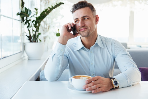 Immagine di uomo adulto felice parlando sul cellulare nero, quando seduto da solo nel piacevole ristorante con una tazza di cappuccino