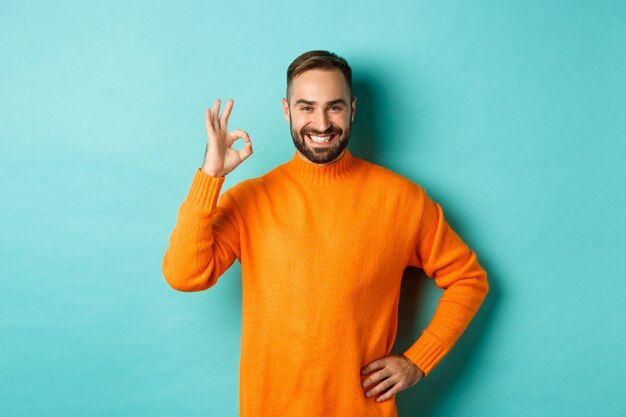 Immagine di uomo bello fiducioso che mostra segno giusto, approva e accetta, come qualcosa di buono, in piedi in maglione arancione.