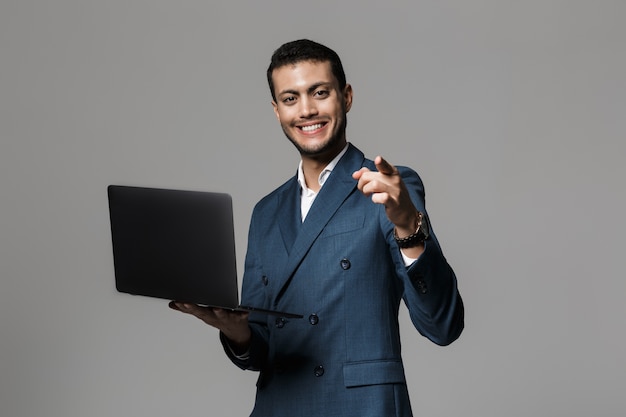 Immagine di uomo d'affari arabo bello anni '30 in abito formale sorridente e tenendo il computer portatile, isolato sopra il muro grigio