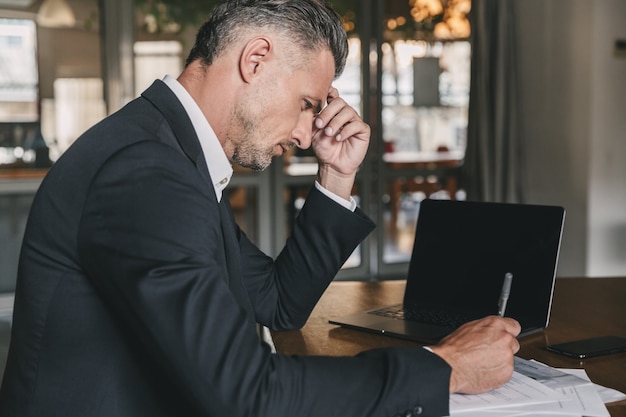 Immagine di uomo d'affari caucasico bello che indossa una camicia bianca e abito nero seduto al tavolo in ufficio, durante il lavoro con documenti e laptop