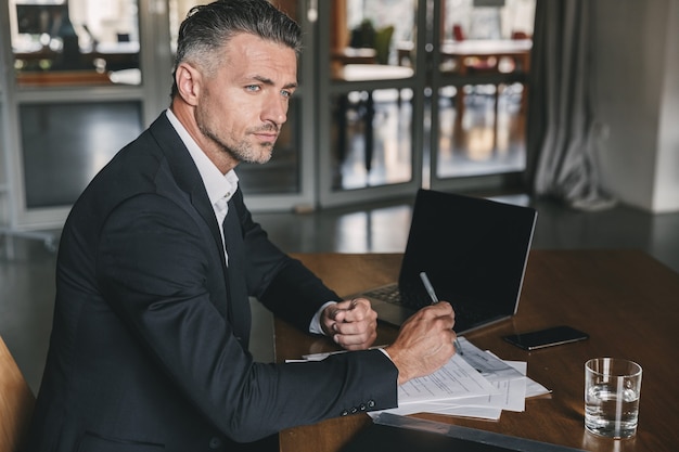 Immagine di uomo d'affari concentrato fiducioso 30s indossa una camicia bianca e abito nero seduto al tavolo in ufficio, durante il lavoro con documenti e laptop