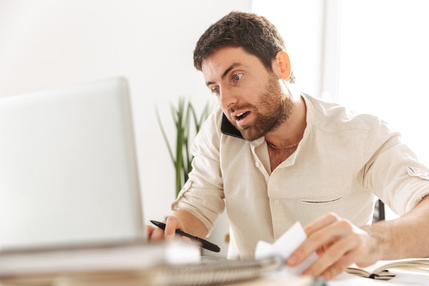 Immagine di uomo d'affari europeo 30s indossa una camicia bianca parlando al telefono cellulare, mentre è seduto a tavola in un ufficio moderno