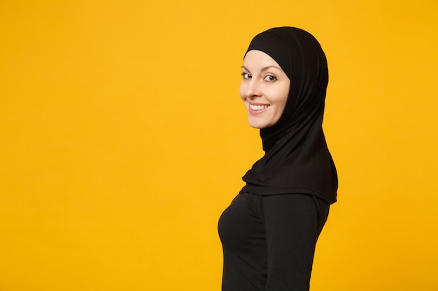 Immagine di vista laterale di bella giovane donna musulmana araba in vestiti neri di hijab che posano isolata sulla parete gialla, ritratto. Concetto di stile di vita dell'Islam religioso della gente.