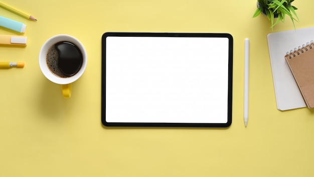 Immagine di vista superiore della compressa ritagliata del computer nero con lo schermo in bianco bianco, tazza di caffè, penna, taccuino, pianta in vaso che mette insieme sullo scrittorio funzionante. Concetto di posto di lavoro ordinato.