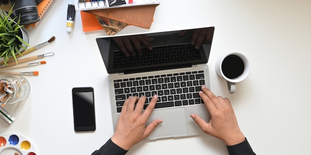 Immagine di vista superiore delle mani del grafico che scrivono sul computer portatile nero dello schermo in bianco che mette sul tavolo di lavoro bianco con la tazza di caffè, il cellulare nero dello schermo in bianco e l'attrezzatura della pittura.