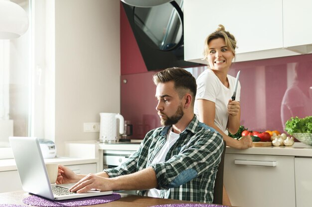 Immagine divertente di un ragazzo con un laptop e la sua ragazza pazza con un coltello in cucina