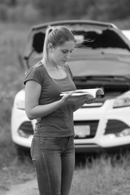 Immagine in bianco e nero di una donna depressa che guarda la sua auto rotta e legge il manuale del proprietario