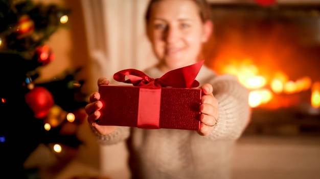Immagine in primo piano di una giovane donna che mostra una scatola regalo di Natale rossa a porte chiuse mentre è seduta accanto all'albero di Natale e al caminetto acceso