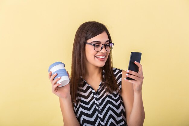 Immagine interna di una giovane donna europea di bell'aspetto isolata sullo sfondo pesca con il caffè in mano e il telefono in mano e sorridendo mentre consulta o legge il messaggio