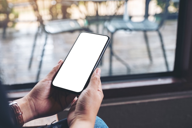 Immagine mockup delle mani di una donna che tengono il telefono cellulare nero con lo schermo del desktop vuoto nel caffè