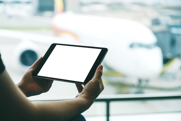 Immagine mockup delle mani di una donna che tiene e usa un tablet pc nero con schermo bianco vuoto mentre è seduta in aeroporto