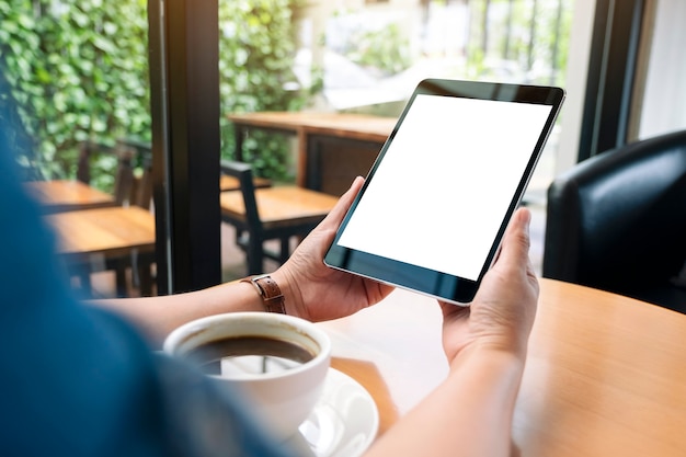 Immagine mockup di mani che tengono un tablet pc nero con schermo bianco vuoto con tazza di caffè sul tavolo di legno