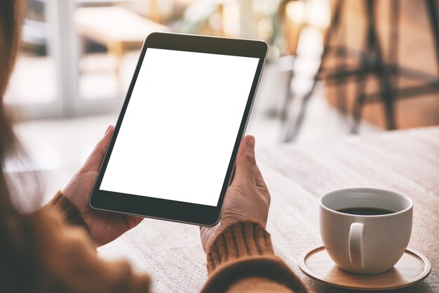 Immagine mockup di mani che tengono un tablet pc nero con schermo bianco vuoto con tazza di caffè sul tavolo di legno
