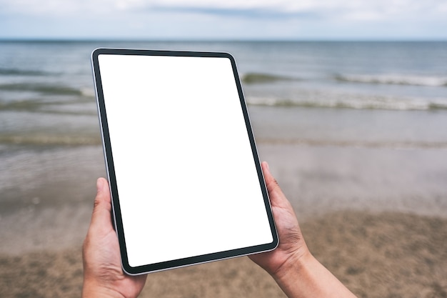 Immagine mockup di mani che tengono un tablet pc nero con schermo desktop vuoto in riva al mare