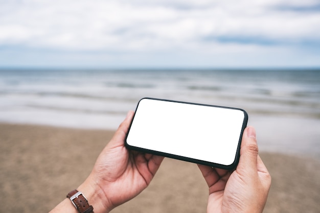 Immagine mockup di mani che tengono un telefono cellulare nero con schermo desktop vuoto in riva al mare