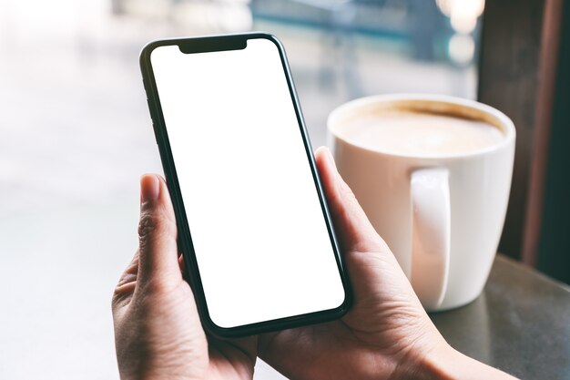 Immagine mockup di mani che tengono un telefono cellulare nero con schermo vuoto con tazza di caffè sul tavolo di legno