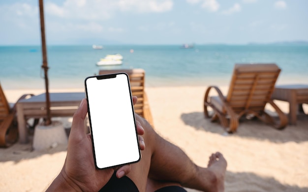 Immagine mockup di un uomo che tiene il telefono cellulare con lo schermo del desktop vuoto mentre è seduto sulla spiaggia