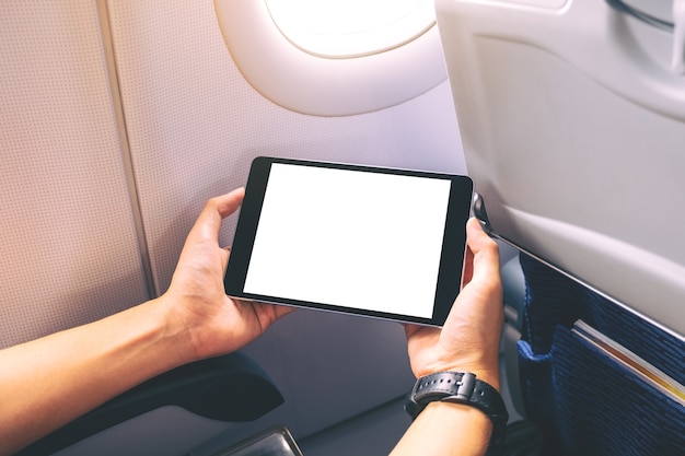 Immagine mockup di un uomo che tiene in mano e guarda un tablet pc nero con uno schermo desktop bianco vuoto accanto alla finestra di un aeroplano
