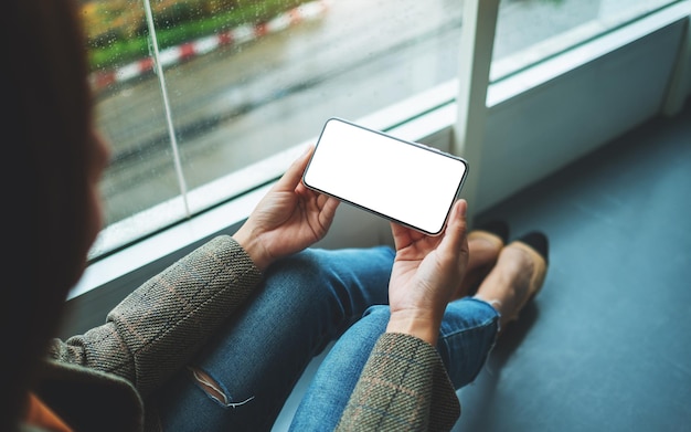 Immagine mockup di una donna che tiene il telefono cellulare con lo schermo del desktop bianco vuoto mentre è seduto sul pavimento