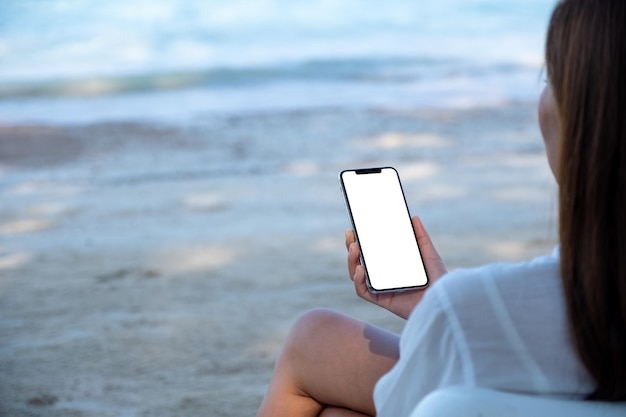 Immagine mockup di una donna che tiene il telefono cellulare con lo schermo del desktop vuoto mentre è seduto sulla spiaggia