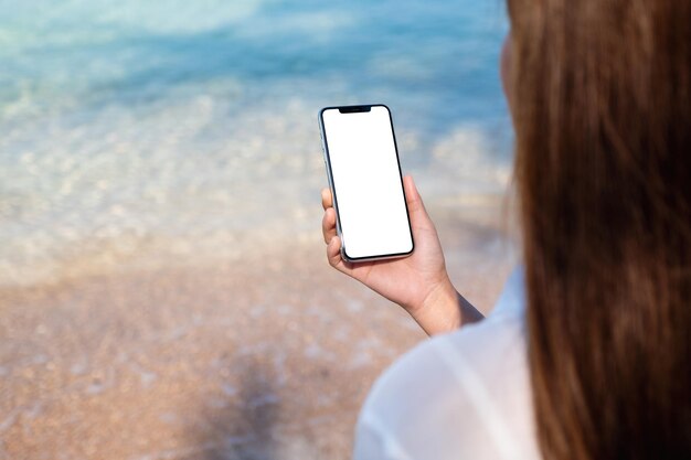 Immagine mockup di una donna che tiene il telefono cellulare con lo schermo del desktop vuoto mentre è seduto sulla spiaggia