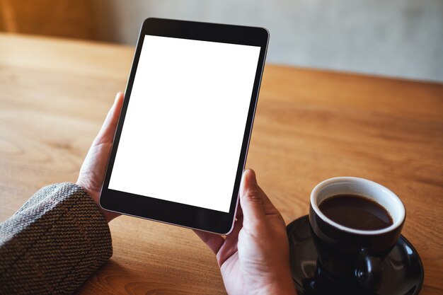 Immagine mockup di una donna che tiene in mano un tablet pc nero con schermo bianco vuoto con una tazza di caffè sul tavolo di legno