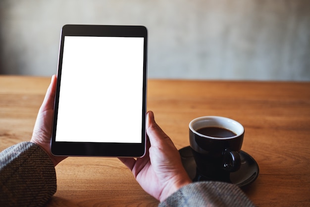 Immagine mockup di una donna che tiene in mano un tablet pc nero con schermo bianco vuoto con una tazza di caffè sul tavolo di legno