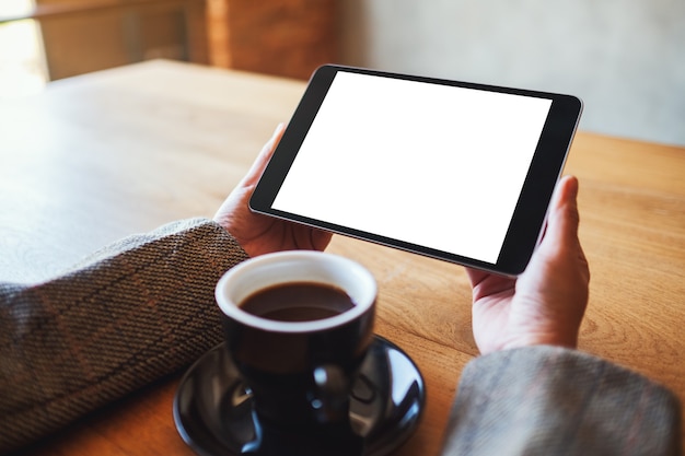 Immagine mockup di una donna che tiene in mano un tablet pc nero con schermo bianco vuoto con una tazza di caffè sul tavolo di legno