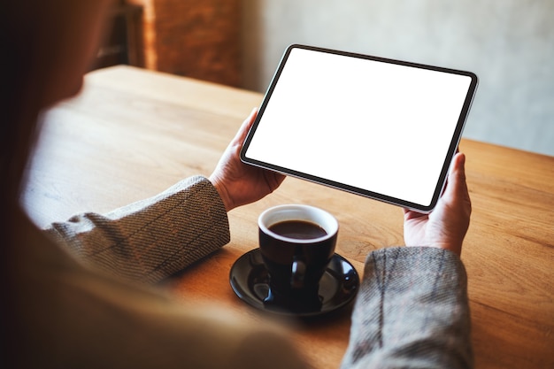 Immagine mockup di una donna che tiene in mano un tablet pc nero con schermo bianco vuoto con una tazza di caffè sul tavolo