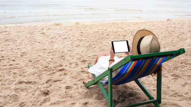 Immagine mockup di una donna che tiene in mano un tablet pc nero con schermo desktop vuoto mentre è sdraiata su una sedia a sdraio