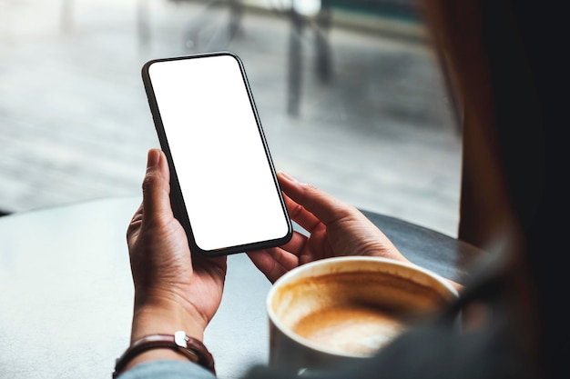 Immagine mockup di una donna che tiene in mano un telefono cellulare con schermo bianco vuoto con una tazza di caffè sul tavolo