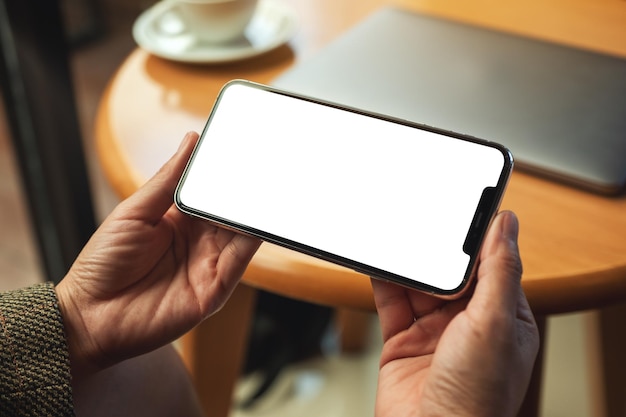 Immagine mockup di una donna che tiene in mano un telefono cellulare con uno schermo desktop bianco vuoto con laptop e tazza di caffè sul tavolo