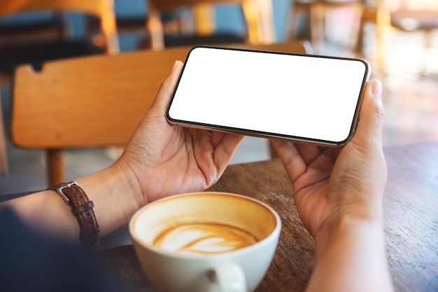 Immagine mockup di una donna che tiene in mano un telefono cellulare nero con schermo vuoto con una tazza di caffè su un tavolo di legno